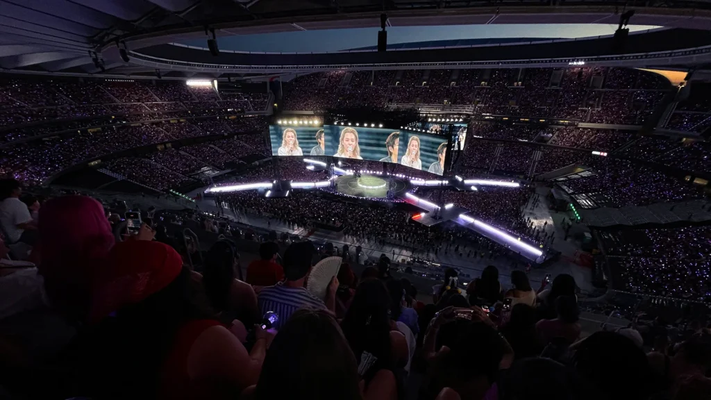 Vista panorámica del Riyadh Air Metropolitano de Madrid durante el concierto de BTS, con el escenario circular y las pasarelas en forma de equis, y el público con lightsticks encendidos.